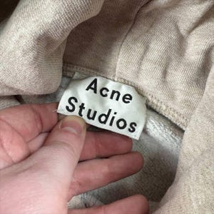K&ouml;nnte beinhalten: Nahaufnahme eines beigefarbenen Hoodies mit dem Etikett "Acne Studios" in schwarzer Schrift. Das Etikett ist wei&szlig; und rechteckig. Der Hoodie besteht aus einem weichen, strukturierten Material.