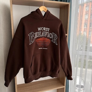 Può includere: Una felpa con cappuccio marrone con il testo "WORST BEHAVIOR" in lettere bianche e rosse. Il testo è circondato da una grafica di un tramonto con strisce rosse e arancioni. Il testo "SOME RISE, SOME REST" è stampato in bianco sotto la grafica.