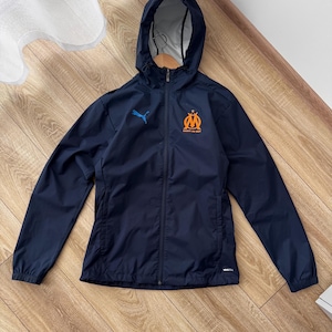 May include: Navy blue hooded jacket with a full-length zip. The jacket has a blue Puma logo and an orange emblem. The jacket is made of a lightweight material. The jacket is suitable for sports or casual wear.