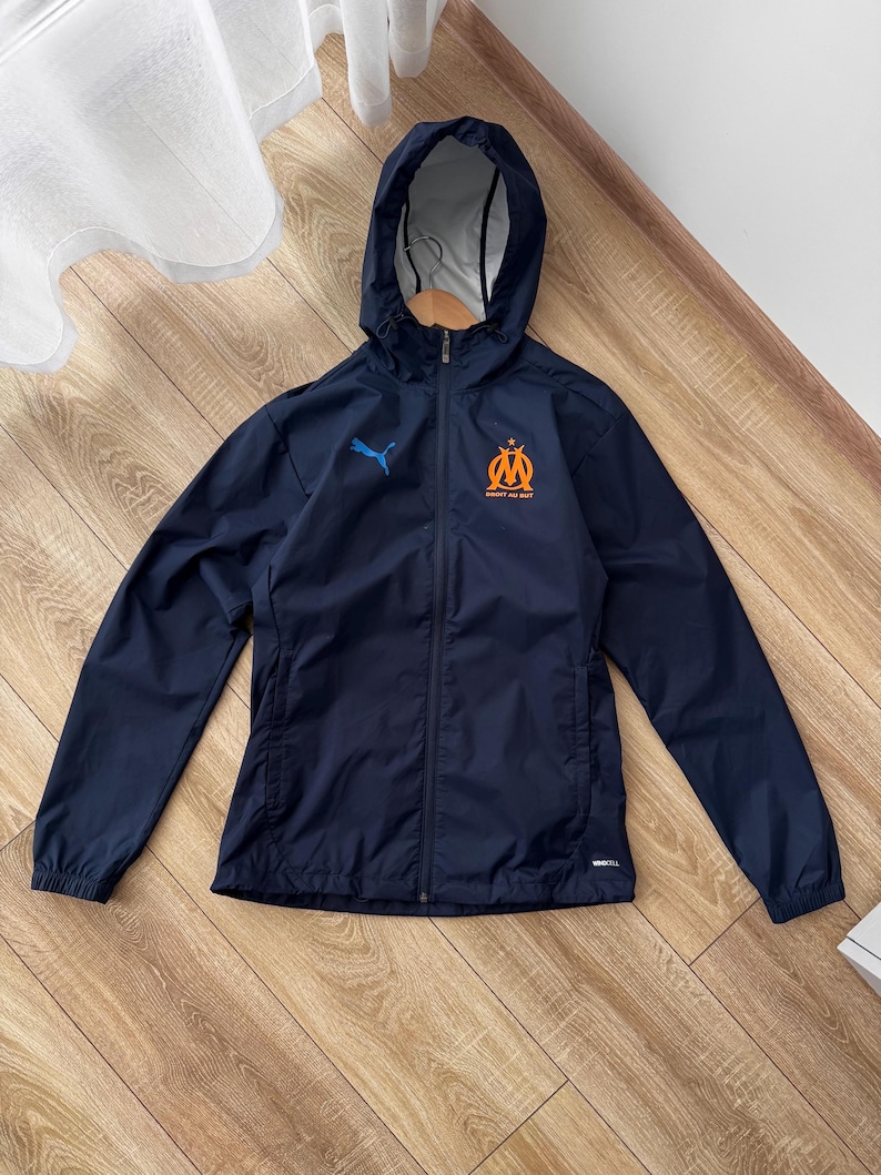 May include: Navy blue hooded jacket with a full-length zip. The jacket has a blue Puma logo and an orange emblem. The jacket is made of a lightweight material. The jacket is suitable for sports or casual wear.
