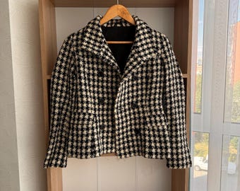 May include: A black and white houndstooth patterned jacket with a double-breasted closure and a collar. The jacket is hanging on a wooden hanger on a shelf.