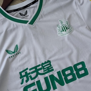 May include: White soccer jersey with green accents, featuring the Newcastle United crest and the Castore logo. The jersey has a V-neck collar and the text "FUN88" in green.