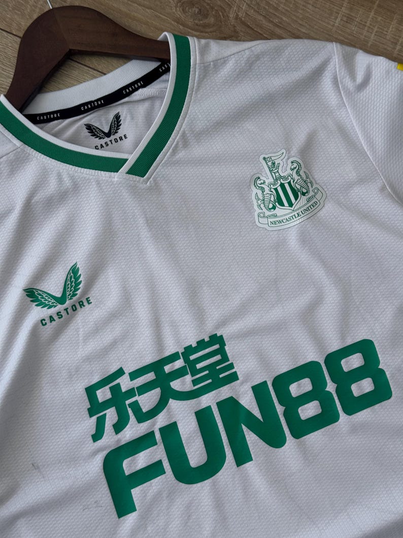 May include: White soccer jersey with green accents, featuring the Newcastle United crest and the Castore logo. The jersey has a V-neck collar and the text "FUN88" in green.