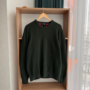 Puede incluir: Un suéter verde oscuro con cuello redondo. El suéter está colgado en una percha de madera en un estante.