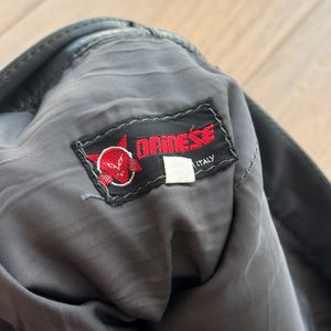 Pu&ograve; includere: Primo piano dell'interno di un capo in pelle nera con fodera grigia. &Egrave; visibile un'etichetta rossa e bianca "DAINESE ITALY", con un logo stilizzato a forma di testa di diavolo. Il capo sembra usurato.