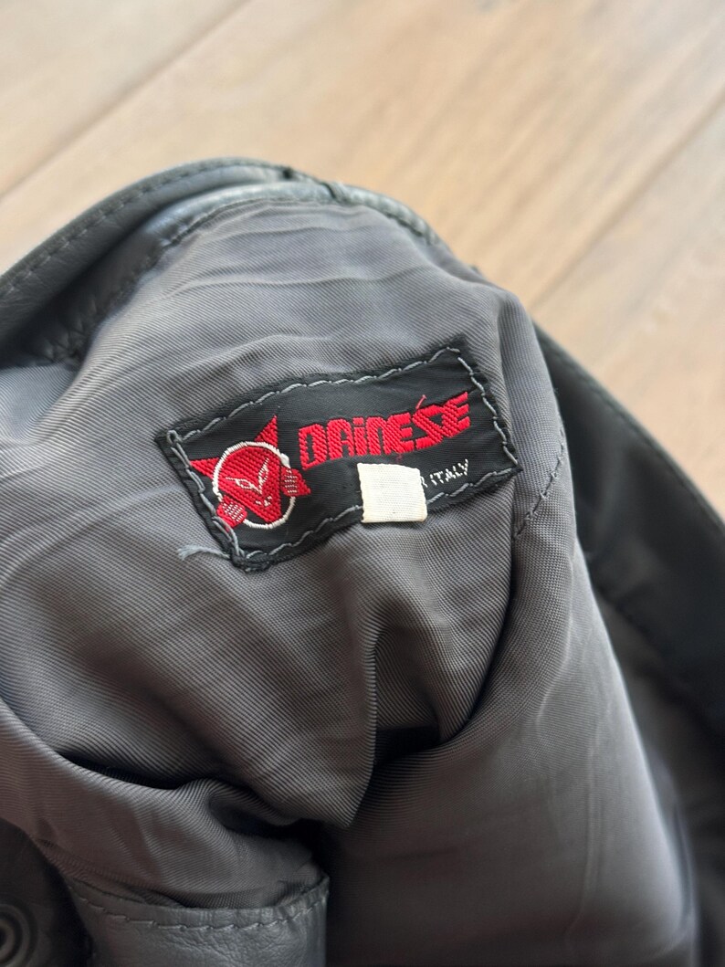 Pu&ograve; includere: Primo piano dell'interno di un capo in pelle nera con fodera grigia. &Egrave; visibile un'etichetta rossa e bianca "DAINESE ITALY", con un logo stilizzato a forma di testa di diavolo. Il capo sembra usurato.