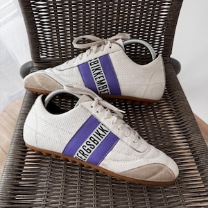 May include: A pair of off-white sneakers with a perforated texture, featuring a prominent purple vertical stripe across the side. The stripe displays the black text BIKKEMBERGS. The shoes have beige suede toe caps, white laces, and a brown rubber sole.