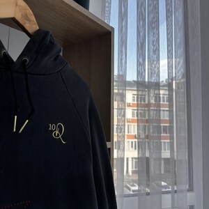 Op de afbeelding: Een zwart hoodie met een geborduurd logo in goud dat "10Q" zegt. De hoodie hangt aan een houten hanger voor een raam met witte gordijnen.