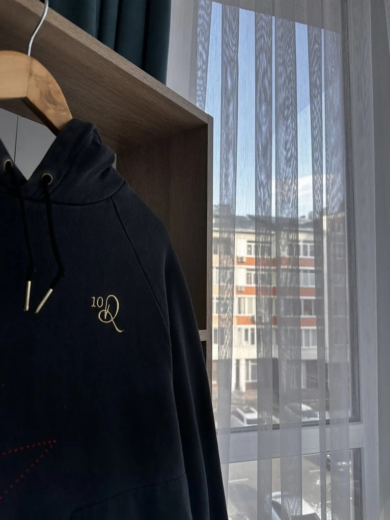 Op de afbeelding: Een zwart hoodie met een geborduurd logo in goud dat "10Q" zegt. De hoodie hangt aan een houten hanger voor een raam met witte gordijnen.