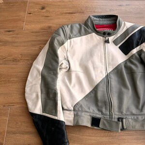 Op de afbeelding: Een wit en grijs leren motorjack met zwarte accenten en een rode rits. Het jack heeft een riem op de taille.