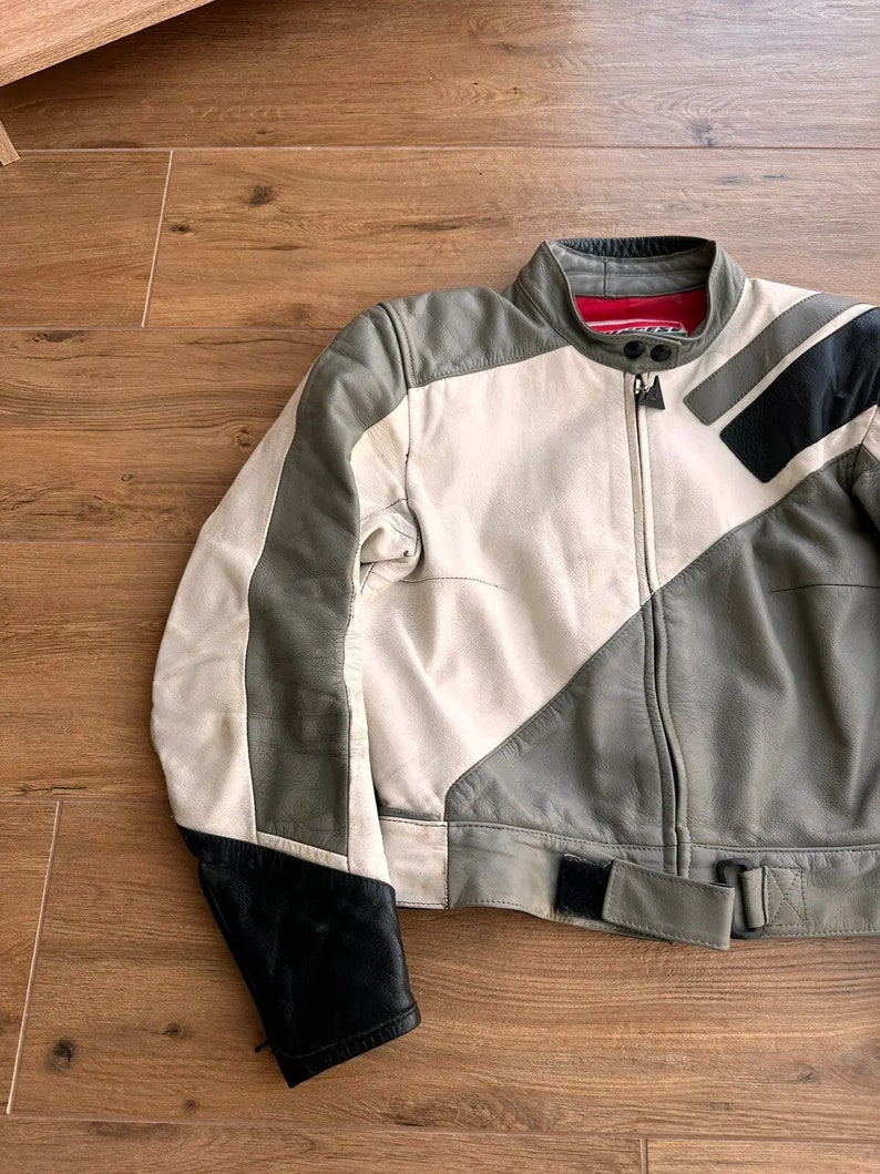 Op de afbeelding: Een wit en grijs leren motorjack met zwarte accenten en een rode rits. Het jack heeft een riem op de taille.