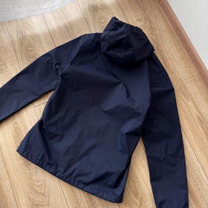 May include: A navy blue hooded jacket with long sleeves. The jacket features a full zip and a hood. The material looks lightweight and water-resistant. The jacket is on a wooden surface.