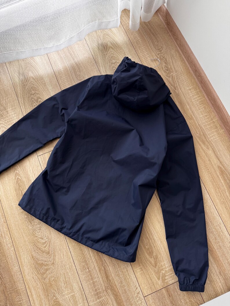 May include: A navy blue hooded jacket with long sleeves. The jacket features a full zip and a hood. The material looks lightweight and water-resistant. The jacket is on a wooden surface.