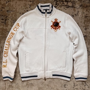 Könnte beinhalten: Cremefarbene Jacke mit Reißverschluss und goldfarbenem Schriftzug "R.L. CHALLENGE CUP" auf dem linken Ärmel. Die Jacke hat ein Wappen auf der Brust und marineblaue und grüne Streifen an Kragen, Bündchen und Saum.