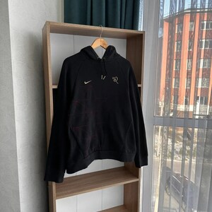 Op de afbeelding: Zwarte hoodie met een rood en wit geometrisch ontwerp op de voorkant. Het Nike-logo bevindt zich op de linkerborst en de tekst "10R" op de rechterborst.