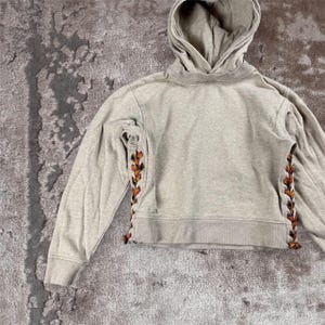 K&ouml;nnte beinhalten: Ein hellgrauer Kapuzenpullover mit langen &Auml;rmeln. Die Seiten der &Auml;rmel haben ein dekoratives Design in Orange, Braun und Schwarz. Der Pullover hat eine Kapuze mit Kordelzug und einen verk&uuml;rzten Saum.
