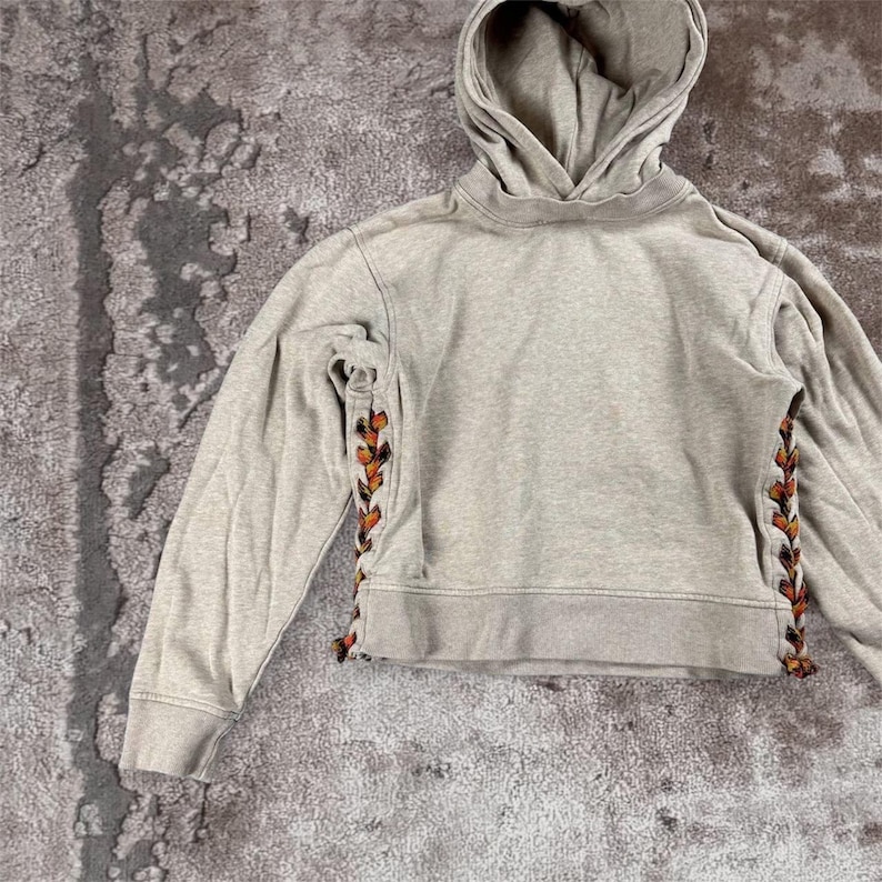 K&ouml;nnte beinhalten: Ein hellgrauer Kapuzenpullover mit langen &Auml;rmeln. Die Seiten der &Auml;rmel haben ein dekoratives Design in Orange, Braun und Schwarz. Der Pullover hat eine Kapuze mit Kordelzug und einen verk&uuml;rzten Saum.