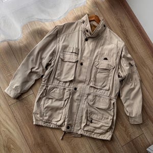 Peut inclure: Une veste utilitaire beige dotée d'un col montant, d'une fermeture à boutons sur le devant et de multiples poches de style cargo sur la poitrine et le bas. La veste comprend des manches longues avec une petite poche zippée sur le bras gauche.