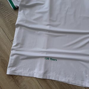 May include: White athletic jersey with green and yellow accents. The jersey has a green collar and sleeve trim. The text "130 Years" is printed in green on the lower right side of the jersey. The jersey is made of a breathable, textured fabric.