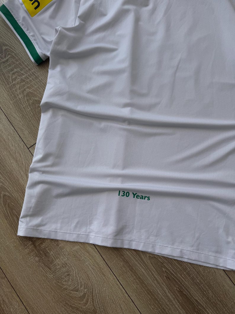 May include: White athletic jersey with green and yellow accents. The jersey has a green collar and sleeve trim. The text "130 Years" is printed in green on the lower right side of the jersey. The jersey is made of a breathable, textured fabric.