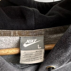Op de afbeelding: Grijze Nike hoodie met een zwarte trekkoord en een wit label met de tekst "NIKE" en "L" met maatgegevens in inches en centimeters. Het label vermeldt ook "Made in Indonesia" en "Fabrique en Indonesie".