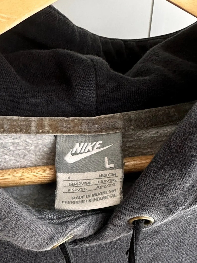 Op de afbeelding: Grijze Nike hoodie met een zwarte trekkoord en een wit label met de tekst "NIKE" en "L" met maatgegevens in inches en centimeters. Het label vermeldt ook "Made in Indonesia" en "Fabrique en Indonesie".