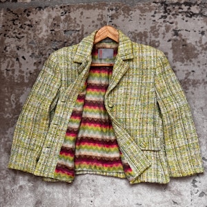 Puede incluir: Una blazer de tweed verde claro con un estampado de cuadros. La blazer tiene un forro a rayas de colores y se muestra en una percha de madera. La chaqueta tiene dos bolsillos delanteros y tres botones.