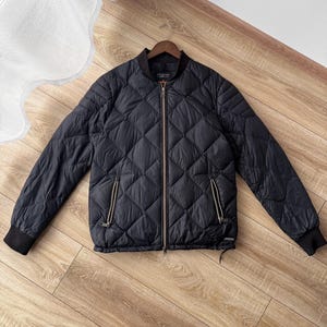 Puede incluir: Chaqueta bomber acolchada negra con un patrón de diamantes. La chaqueta tiene una cremallera frontal, dos bolsillos laterales y puños acanalados. La chaqueta se muestra sobre una superficie de madera.