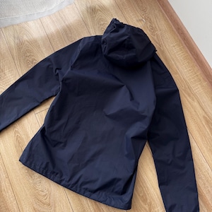 May include: A navy blue hooded jacket with long sleeves. The jacket is made of a lightweight material and has a drawstring hood. The jacket is lying on a wooden floor.