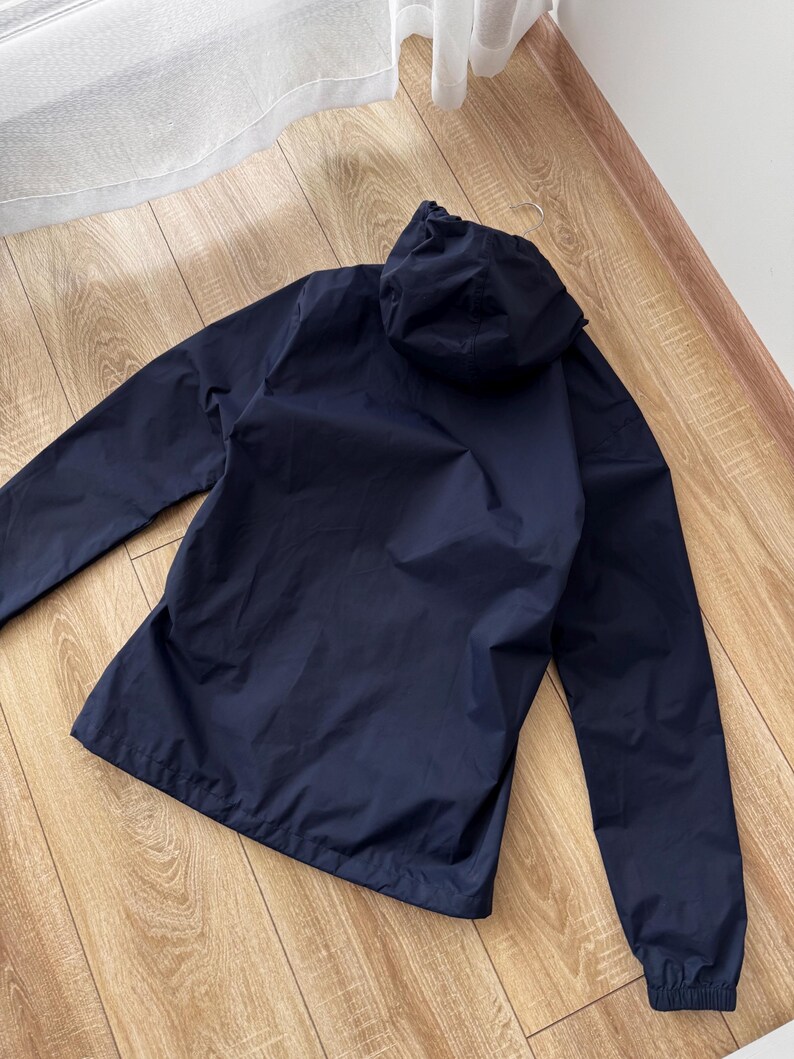 May include: A navy blue hooded jacket with long sleeves. The jacket is made of a lightweight material and has a drawstring hood. The jacket is lying on a wooden floor.