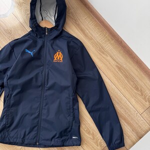 May include: Navy blue hooded jacket with a full-length zip. The jacket features a blue Puma logo on the left chest and an orange and blue emblem on the right chest. It has a hood and two side pockets.