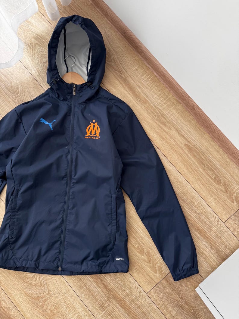 May include: Navy blue hooded jacket with a full-length zip. The jacket features a blue Puma logo on the left chest and an orange and blue emblem on the right chest. It has a hood and two side pockets.