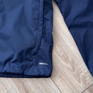 May include: Close-up of a navy blue jacket with a pocket and the word "WINDCELL" printed in white. The jacket is made of a smooth, water-resistant material.