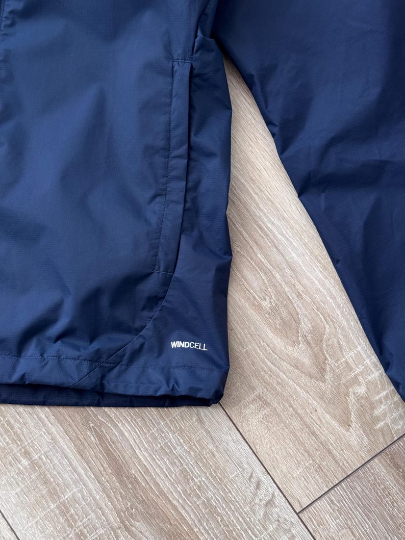 May include: Close-up of a navy blue jacket with a pocket and the word "WINDCELL" printed in white. The jacket is made of a smooth, water-resistant material.