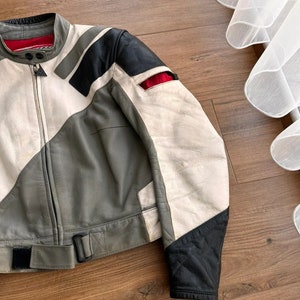 Op de afbeelding: Een grijs, wit en zwart leren motorjack met een rode accent. Het jack heeft een ritssluiting en een riem.