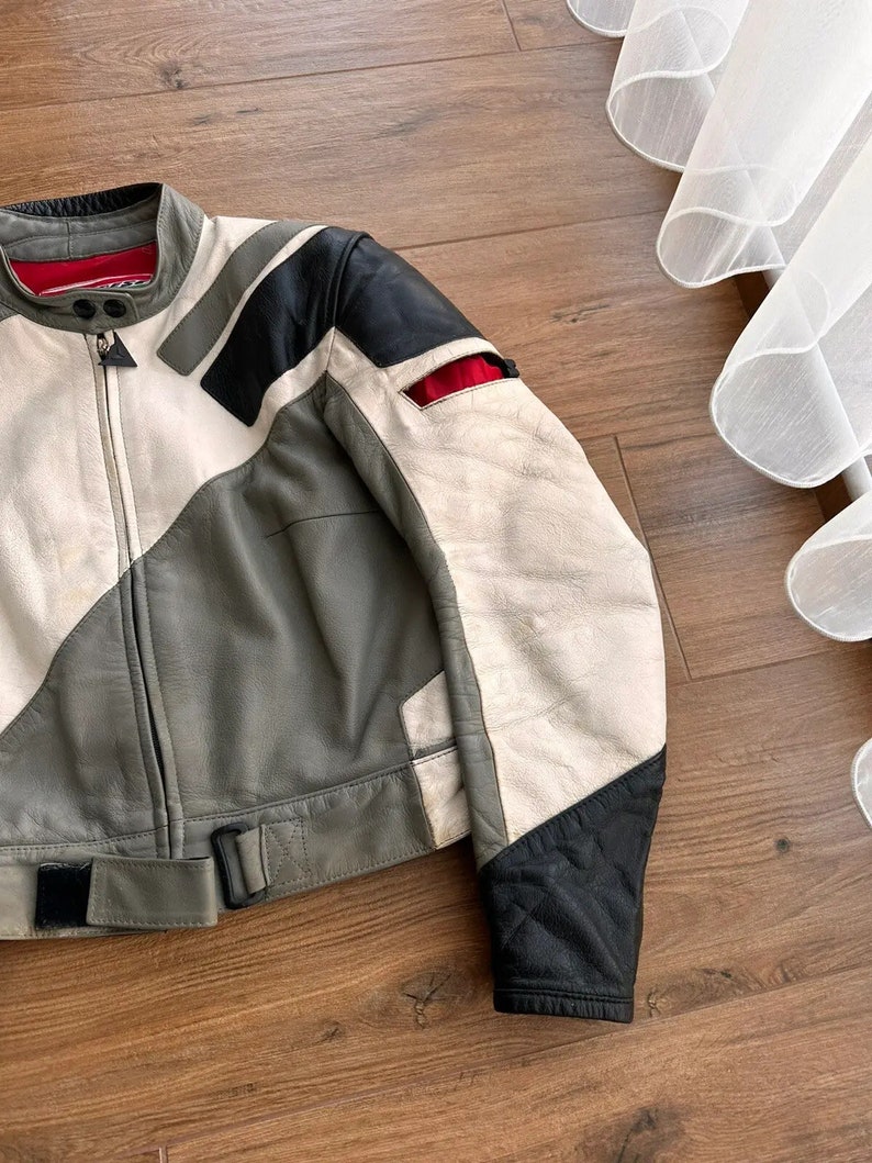 Op de afbeelding: Een grijs, wit en zwart leren motorjack met een rode accent. Het jack heeft een ritssluiting en een riem.