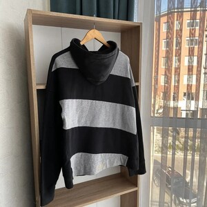 Op de afbeelding: Zwart-grijze gestreepte hoodie met een capuchon. De hoodie hangt aan een houten plank.