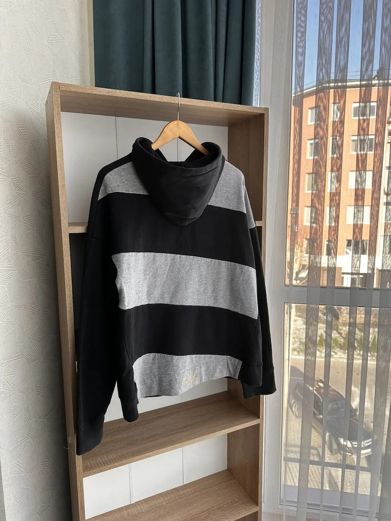 Op de afbeelding: Zwart-grijze gestreepte hoodie met een capuchon. De hoodie hangt aan een houten plank.