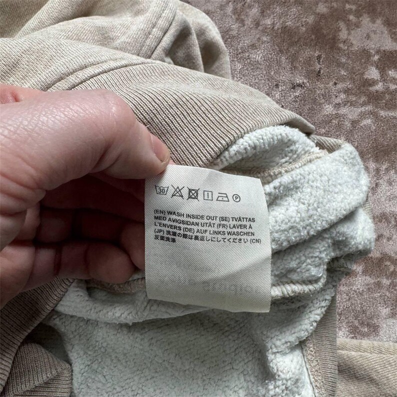 K&ouml;nnte beinhalten: Nahaufnahme eines beigefarbenen Sweatshirts mit wei&szlig;em Innenfutter. Ein Etikett mit Waschanweisungen ist sichtbar, einschlie&szlig;lich Symbolen f&uuml;r Waschen bei 30 Grad Celsius, kein Bleichen und kein B&uuml;geln. Der Text ist in mehreren Sprachen.