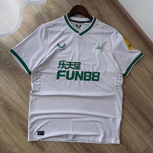 May include: White soccer jersey with green trim and accents. The jersey features the Castore logo, the Newcastle United crest, and the Fun88 sponsor logo in green. The jersey is hanging on a wooden hanger.