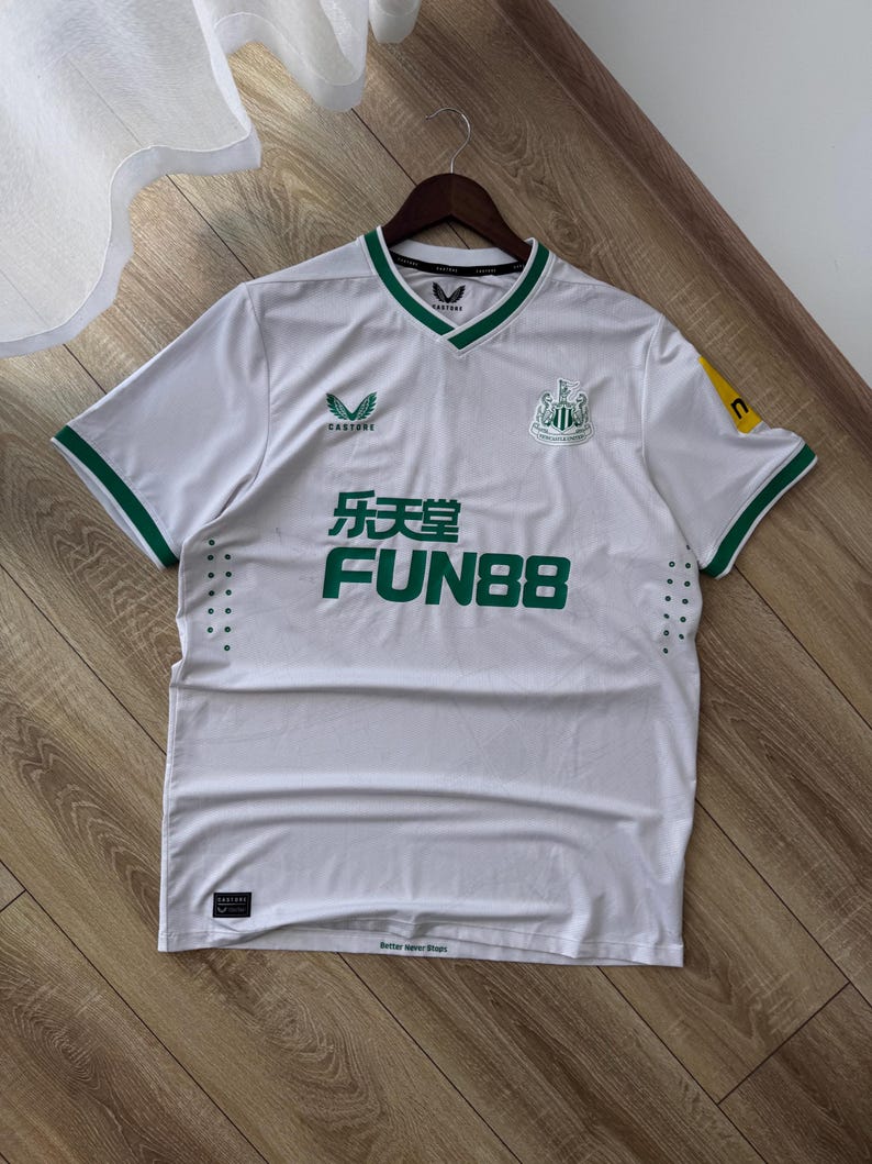 May include: White soccer jersey with green trim and accents. The jersey features the Castore logo, the Newcastle United crest, and the Fun88 sponsor logo in green. The jersey is hanging on a wooden hanger.