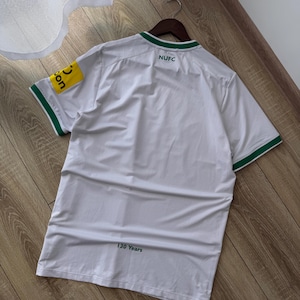 May include: White athletic jersey with green trim on the collar and sleeves. The jersey has the text "NUFC" on the back and a yellow patch with text on the left sleeve. The text "130 Years" is printed at the bottom.