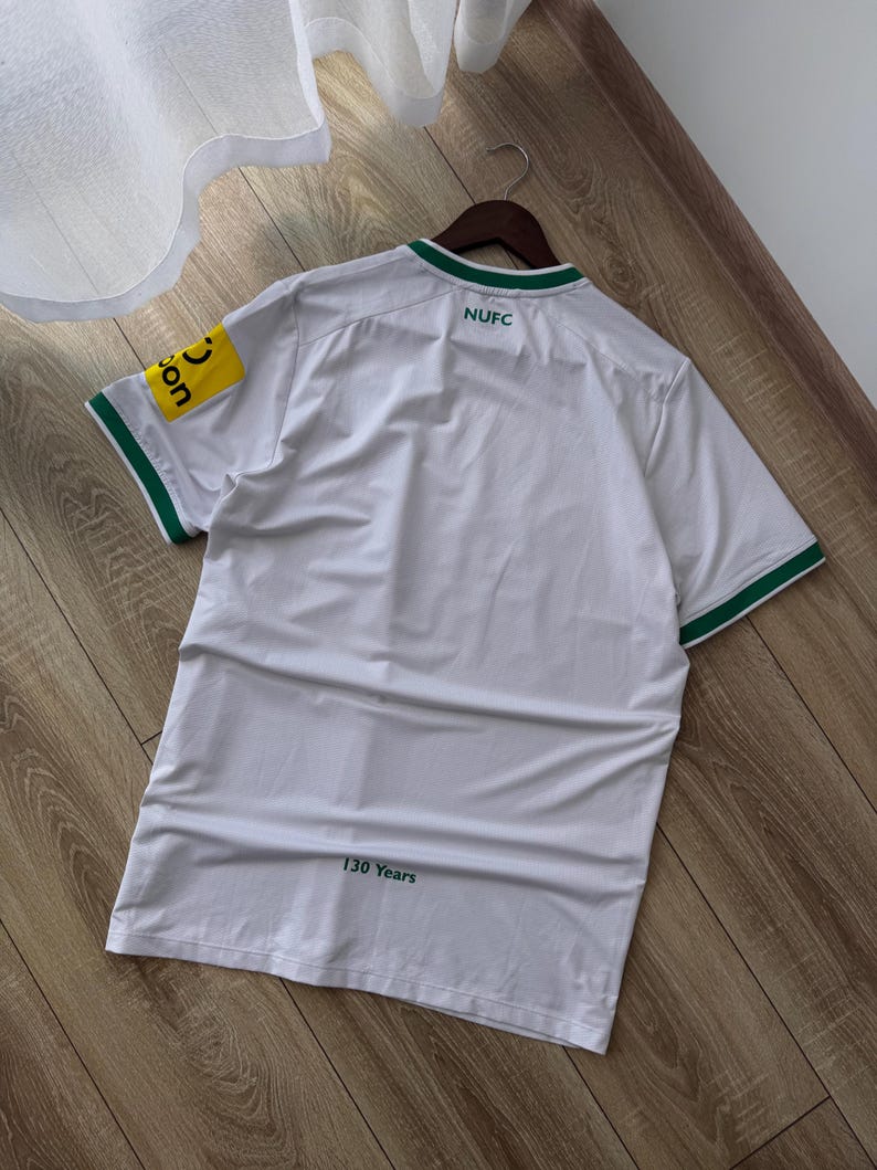 May include: White athletic jersey with green trim on the collar and sleeves. The jersey has the text "NUFC" on the back and a yellow patch with text on the left sleeve. The text "130 Years" is printed at the bottom.