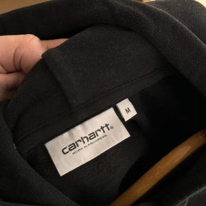 Pu&ograve; includere: Felpa con cappuccio nera Carhartt con un'etichetta bianca che dice "Carhartt" e "Work in Progress" in maiuscolo. L'etichetta ha anche una taglia "M" stampata.
