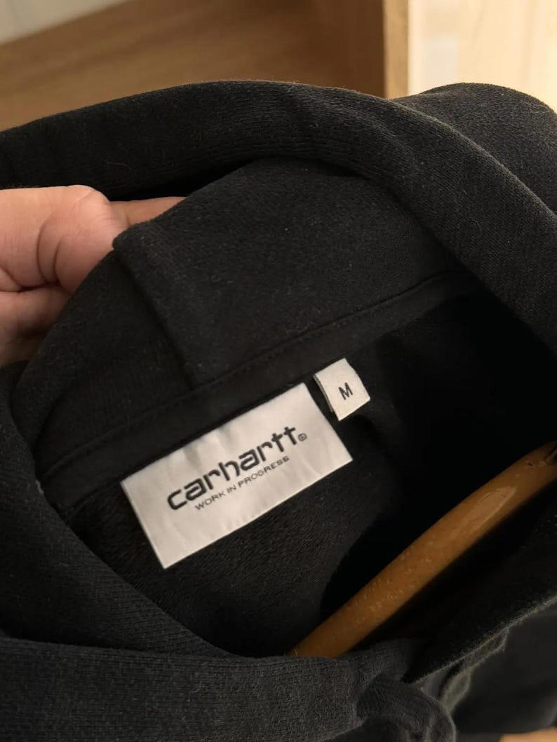 Pu&ograve; includere: Felpa con cappuccio nera Carhartt con un'etichetta bianca che dice "Carhartt" e "Work in Progress" in maiuscolo. L'etichetta ha anche una taglia "M" stampata.