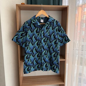 May include: A blue and green short-sleeved button-up shirt with a flame pattern. The shirt is hanging on a wooden shelf.