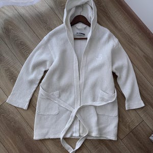 Pode incluir: Um roupão branco em tecido waffle com capuz, mangas compridas, dois bolsos frontais e um cinto de amarrar a condizer. A peça inclui uma etiqueta da marca Jil Sander no decote interior e um pequeno logótipo bordado no lado esquerdo do peito.