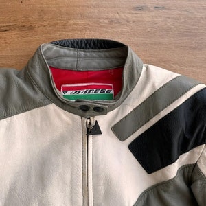 Op de afbeelding: Een grijs, wit en zwart leren motorjack met een ritssluiting en een Dainese-logo aan de linkerkant van de borst.