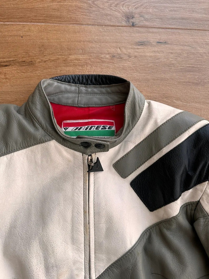 Op de afbeelding: Een grijs, wit en zwart leren motorjack met een ritssluiting en een Dainese-logo aan de linkerkant van de borst.