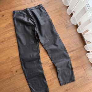 Pu&ograve; includere: Pantaloni in pelle nera dal design classico. I pantaloni hanno un taglio dritto e un'estetica semplice e pulita. La pelle sembra morbida e ha una leggera lucentezza. I pantaloni sono appoggiati su una superficie di legno.